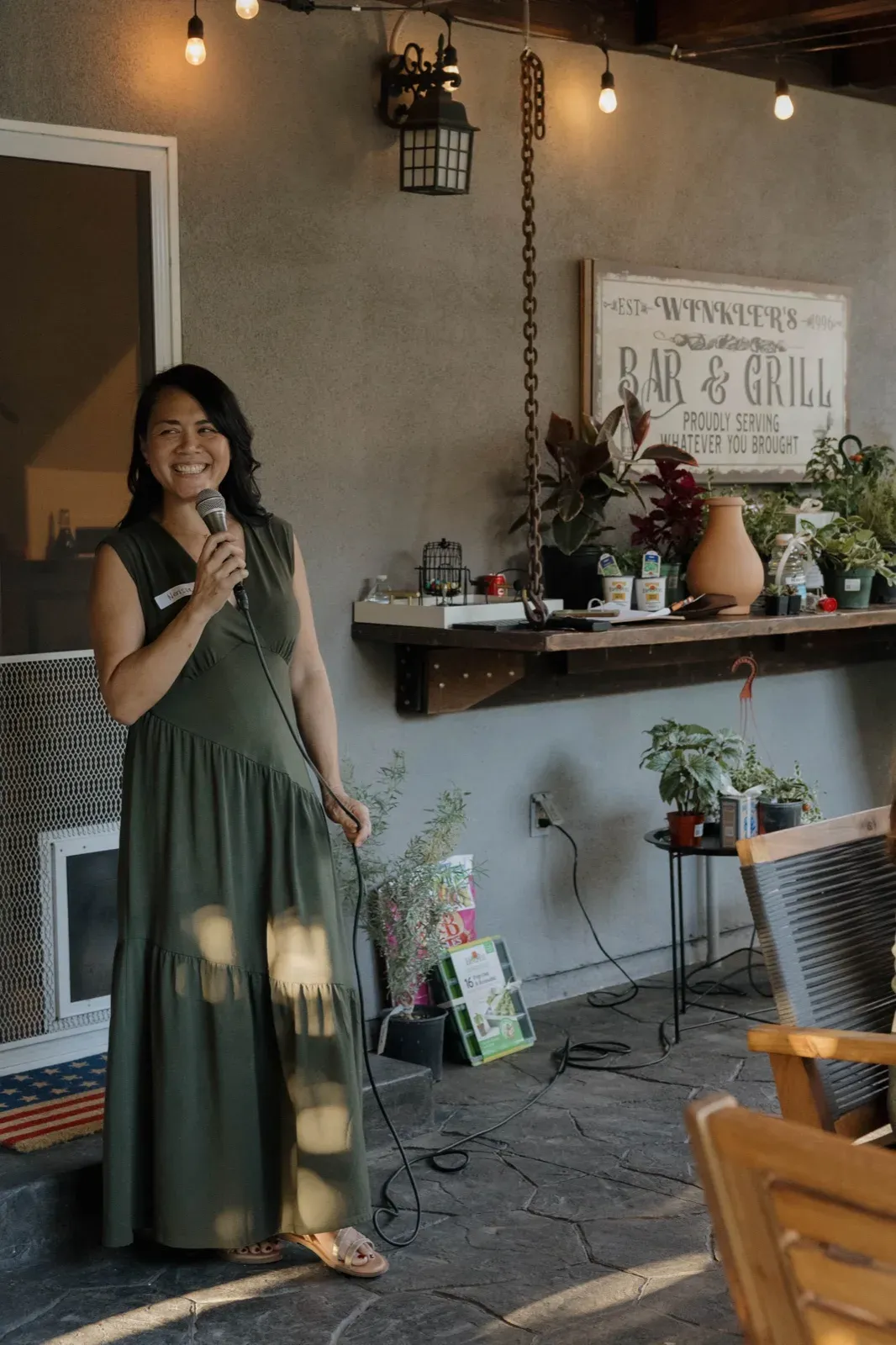 Nerissa hosting a Plant Bingo night, microphone in hand, a shelf of potted plant prizes behind her